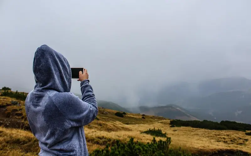 Person in a hood, seen from behind, filming a landscape with a phone